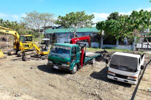 A green Isuzu flatbed crane truck and a white dump truck parked at a job site, with an excavator in the background