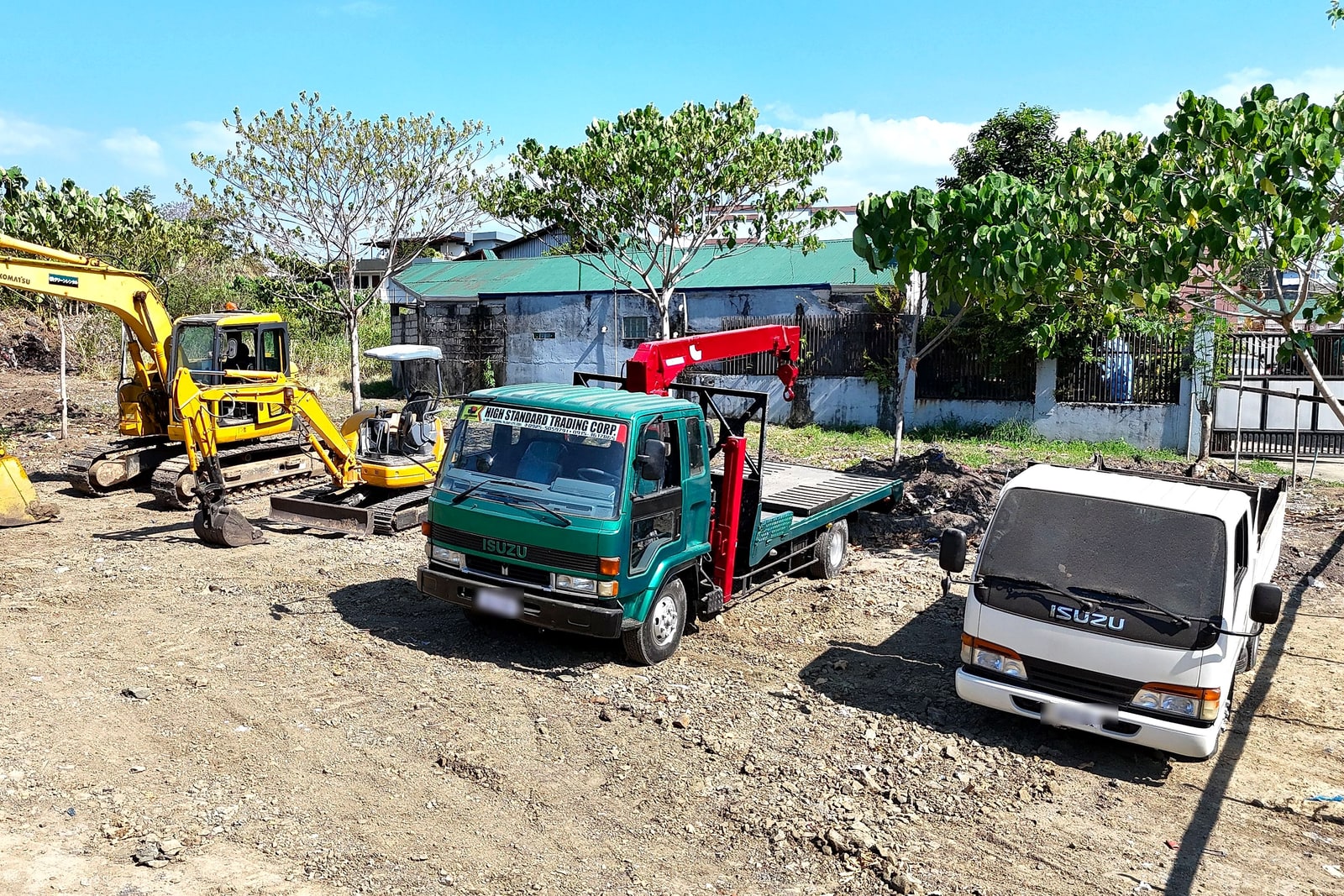 A green Isuzu flatbed crane truck and a white dump truck parked at a job site, with an excavator in the background