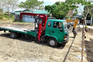 A green boom truck with crane arm extended, parked beside a mini excavator