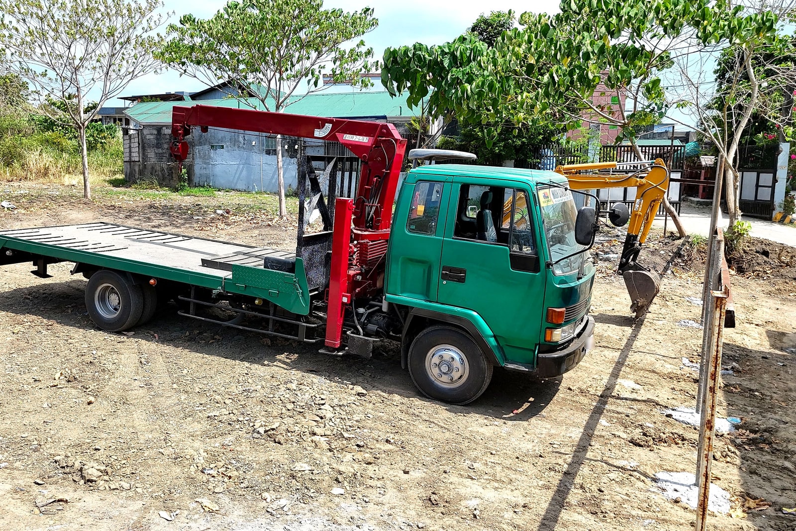 A green boom truck with crane arm extended, parked beside a mini excavator